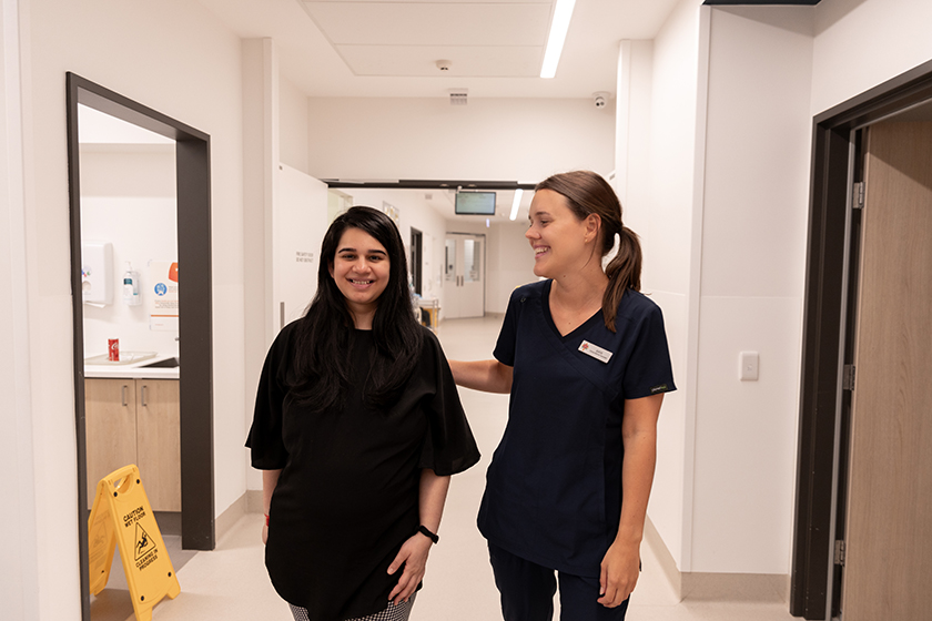 A person walks down a hospital hallway with a caregiver next to them, both are smiling.